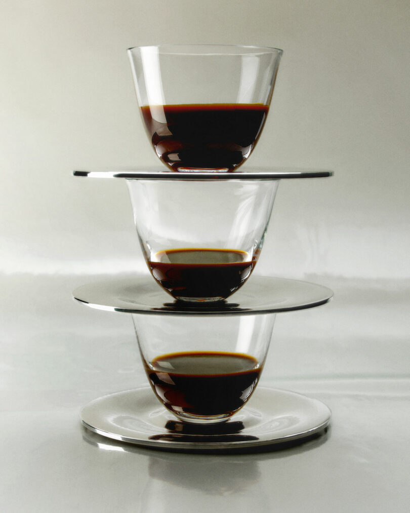Three glass cups from the Artisans Series, partially filled with dark liquid, are stacked vertically and separated by reflective saucers against a plain background.