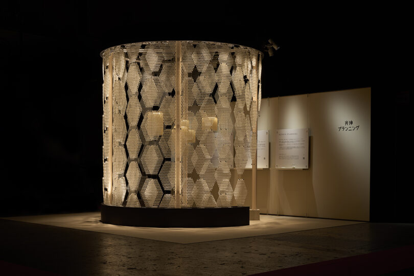 Large cylindrical structure with a honeycomb-patterned exterior, illuminated and displayed in a dimly lit exhibition space. Informational panels on the wall reveal its connection to The Great Tree.