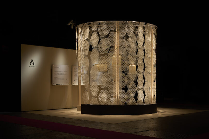 A cylindrical structure called The Great Tree, with a honeycomb-patterned, translucent surface, is illuminated from within and stands on a black base against a dark background with beige display panels.