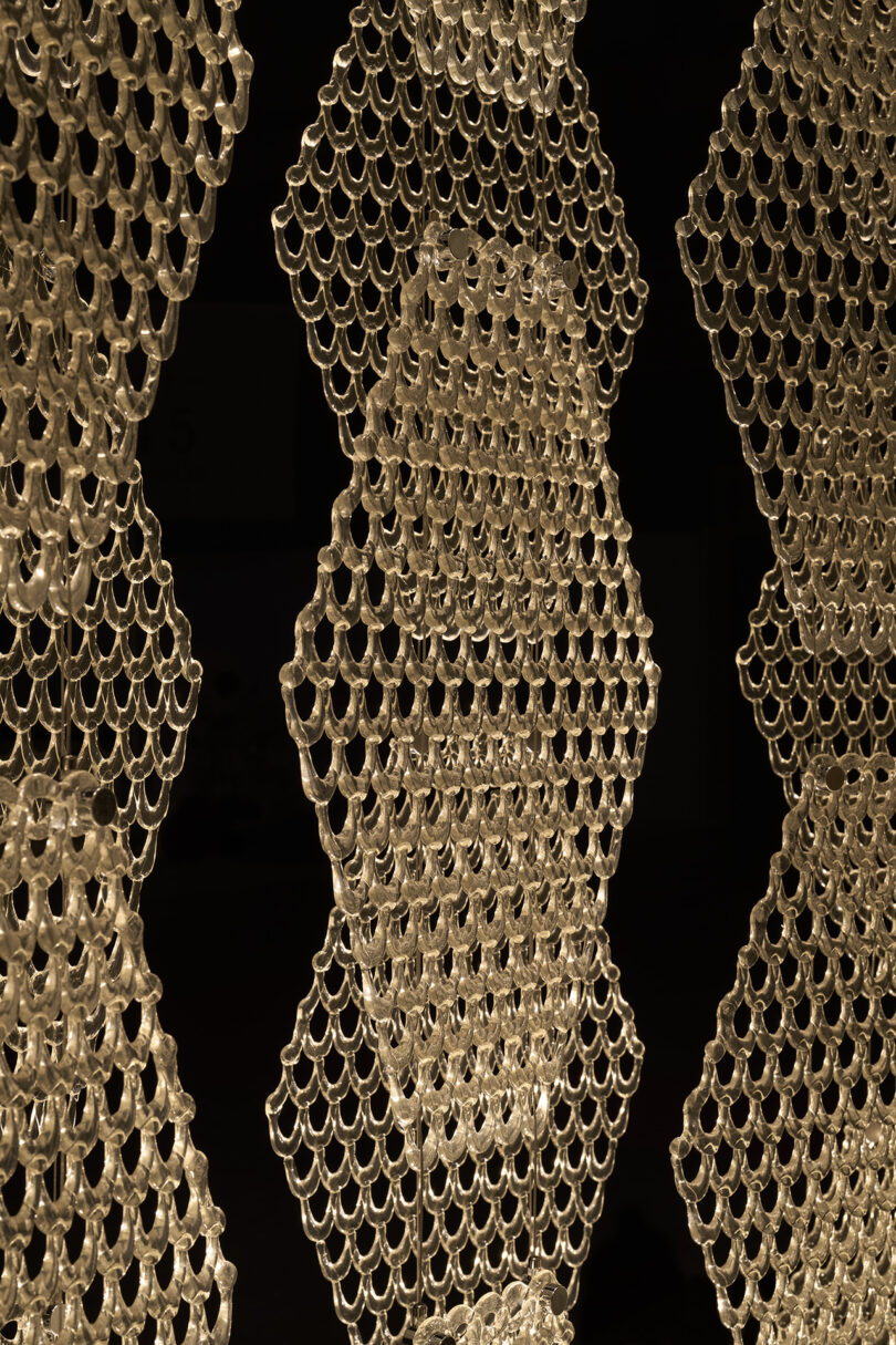 Hexagonal panels made of interlinked translucent rings hang vertically against a black background, creating a geometric pattern reminiscent of The Great Tree’s intricate branches.