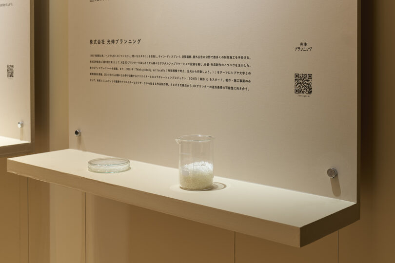 A display shelf with a glass petri dish and a beaker of white granules, set against a light-colored wall featuring Japanese text, a QR code, and an illustration of The Great Tree.