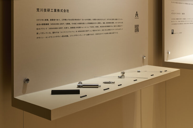 Display case with various minimalist metal objects arranged on a white shelf, set against a beige wall. Above, Japanese text and company information reference The Great Tree, blending tradition with modern design.