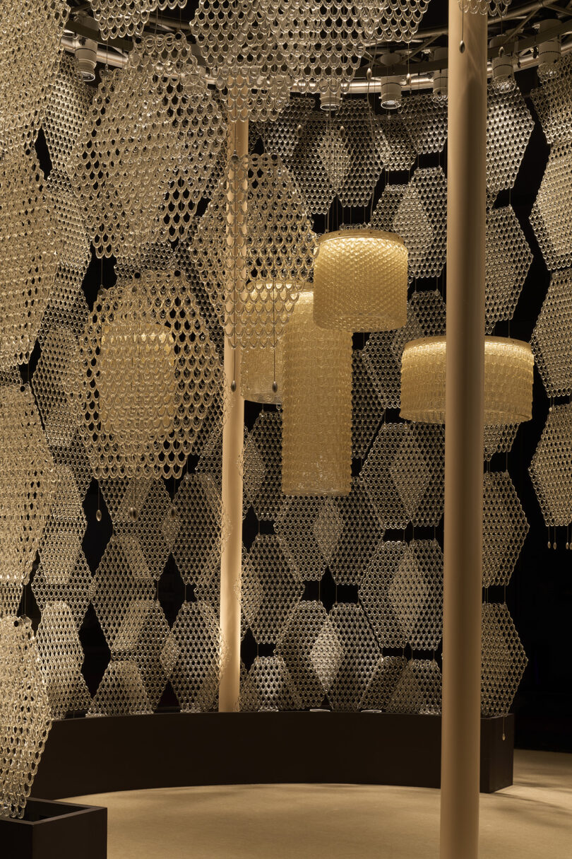 Hexagonal panels and cylindrical light fixtures, evoking the branches of The Great Tree, hang from interlocking, translucent rings in a stunning geometric installation with a dark background and curved seating.