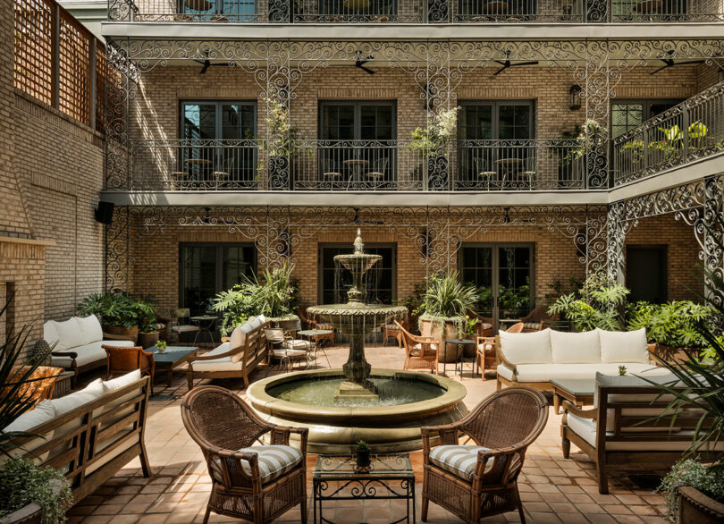 Outdoor courtyard with wrought iron balconies, a central stone fountain, wicker and wooden seating, potted plants, and brick walls, creating a relaxed and elegant atmosphere.