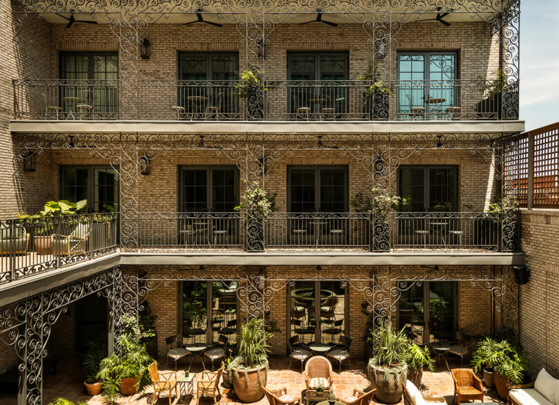 A three-story brick building with ornate iron balconies, potted plants, and outdoor seating in a courtyard.