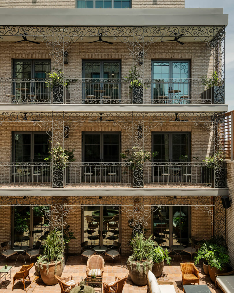 Three-story building with wrought iron balconies, brick facade, large windows, and a ground floor patio area furnished with wicker chairs and surrounded by potted plants.