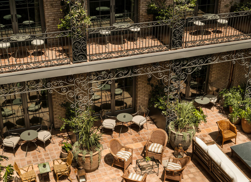 Outdoor courtyard with wrought iron balconies, round tables, wicker chairs, potted plants, and terracotta tile flooring, adjacent to a brick building with large windows.