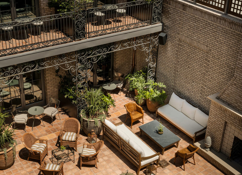 A sunlit courtyard with wicker chairs, cushioned sofas, potted plants, and wrought iron balcony railings against a brick wall.