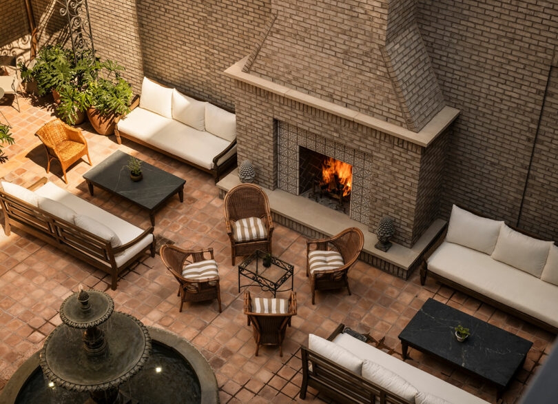Outdoor patio with cushioned seating, wicker chairs around a small table, a lit brick fireplace, potted plants, and a water fountain in the foreground.
