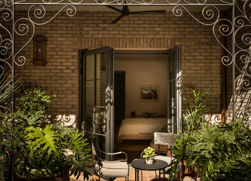 View of a bedroom through open double doors from a patio with potted plants, chairs, and a small table; brick wall and decorative metalwork overhead.
