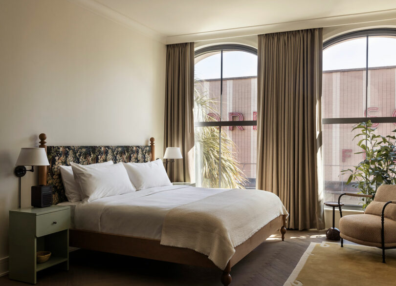 A neatly made bed with white linens sits between two nightstands in a softly lit bedroom with large arched windows, beige curtains, and indoor plants.