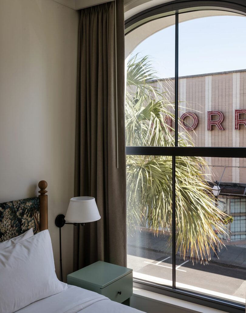 A hotel room with a bed, lamp, and nightstand next to a large arched window showing a palm tree and part of a building with red signage outside.