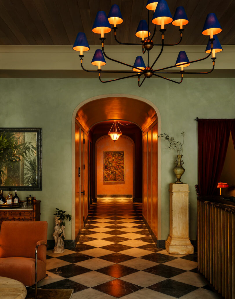 A hallway with checkered floor tiles, warm lighting, green walls, a modern chandelier, and an archway leading to a painting at the end of the corridor.