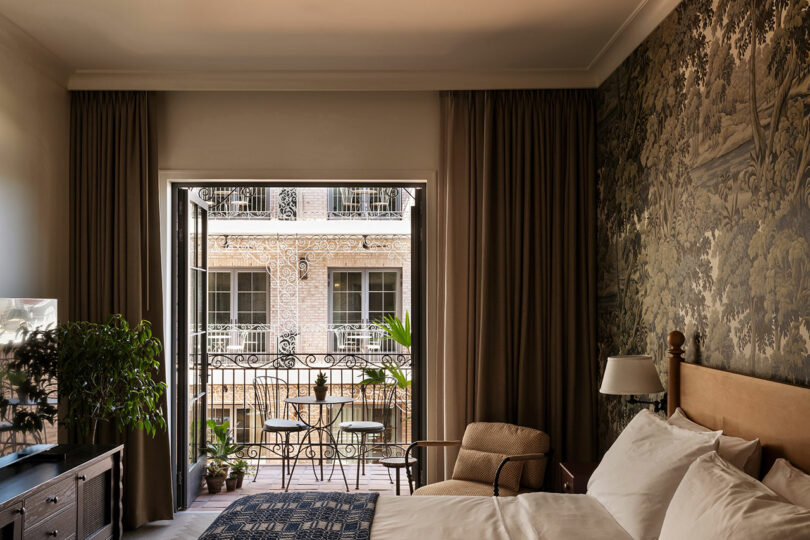 A bedroom with a double bed, armchair, and dresser opens to a balcony with two chairs and a table, overlooking a building with wrought iron railings and windows.
