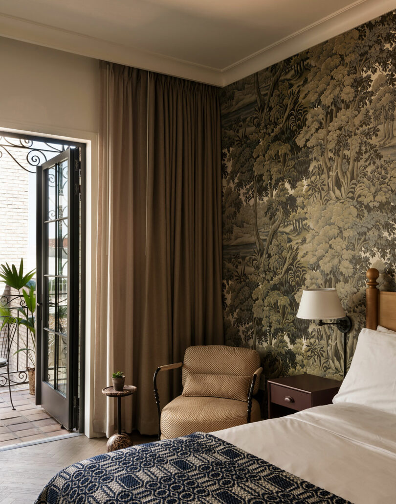 A cozy bedroom with patterned wallpaper, an upholstered chair, a bed with white linens, a small side table, and an open door leading to a brick patio with potted plants.