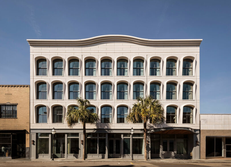 A four-story building with a white facade, large arched windows, and two palm trees in front, set against a clear blue sky.