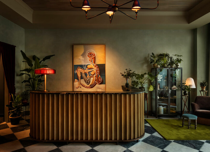 A stylish reception desk with a pleated front, indoor plants, a skeleton artwork, vintage lighting, and a glass display cabinet in a warmly lit room with checkered flooring.