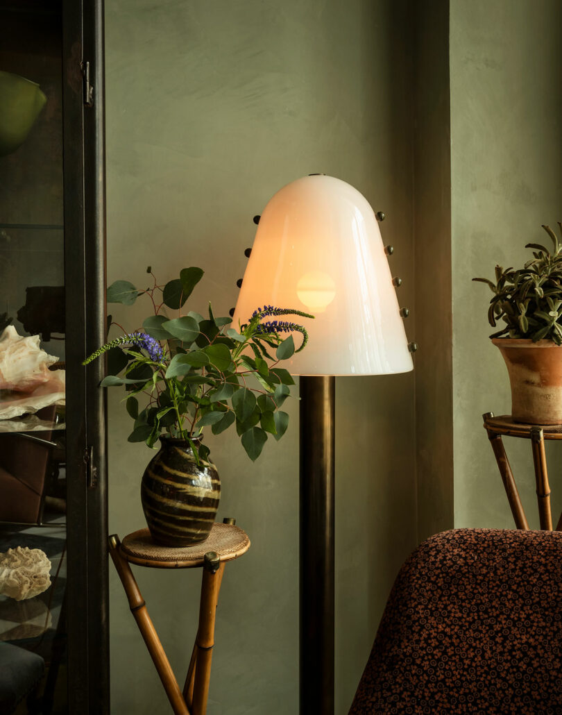 A white dome-shaped floor lamp stands next to a potted plant on a small table; another plant sits in a pot nearby against a green wall.