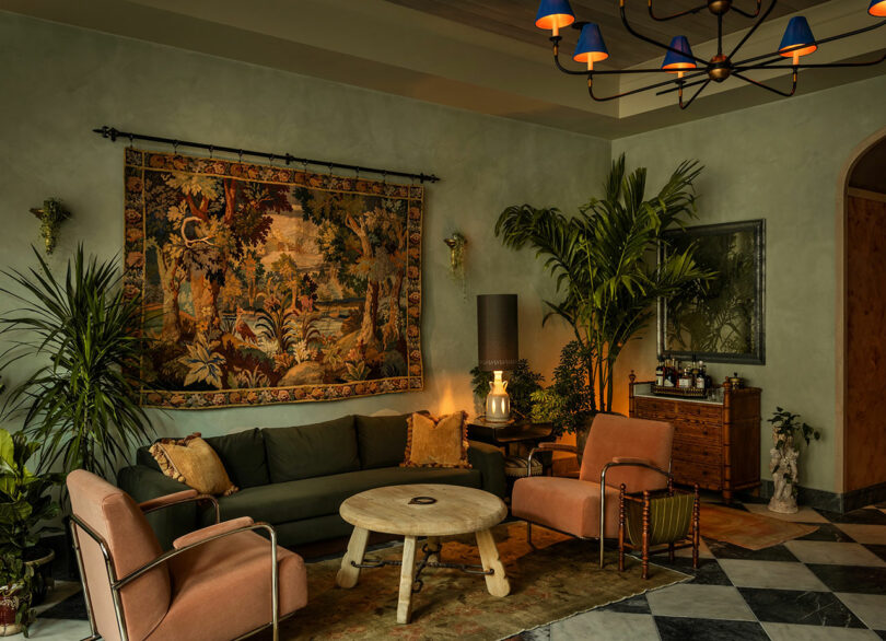 A living room with a green sofa, two pink chairs, a round wooden coffee table, large potted plants, a tapestry on the wall, and a chandelier overhead.