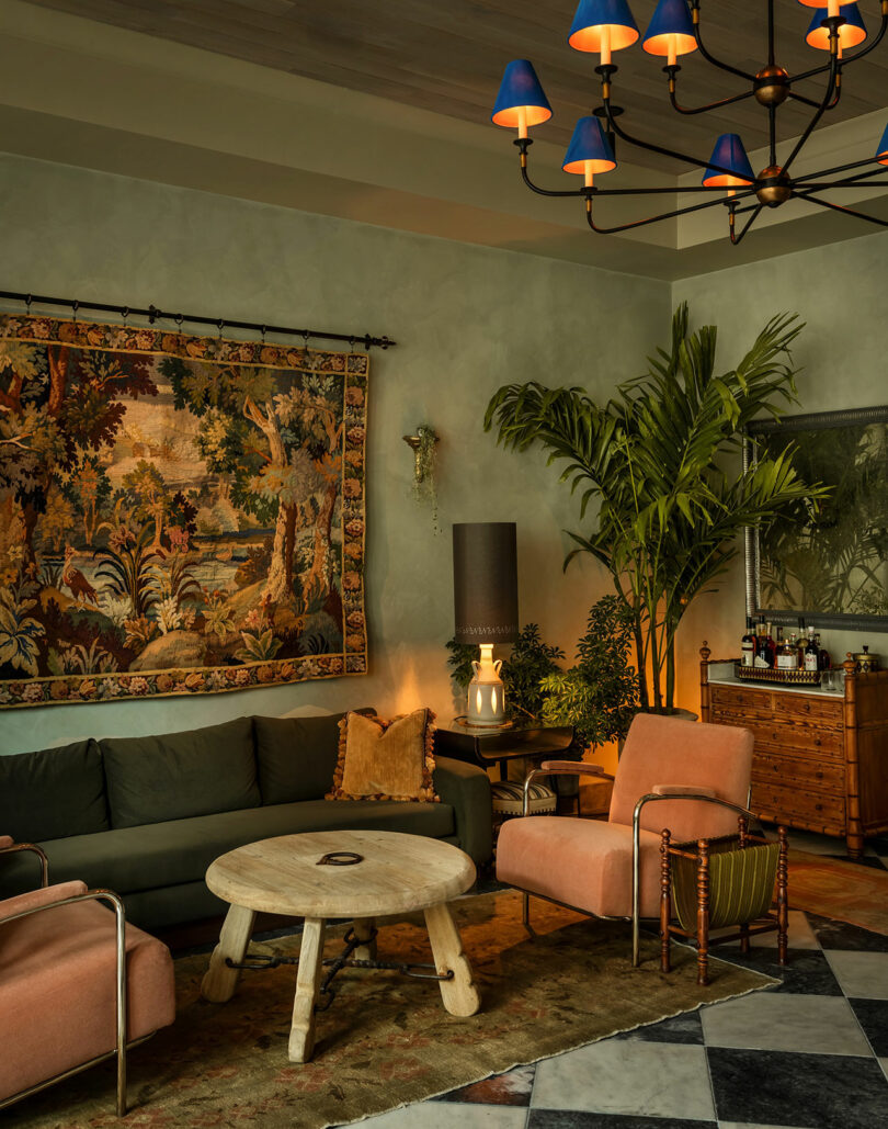 Living room with a tapestry on the wall, green sofa, two pink armchairs, round wooden coffee table, indoor plants, and a chandelier with blue lampshades.