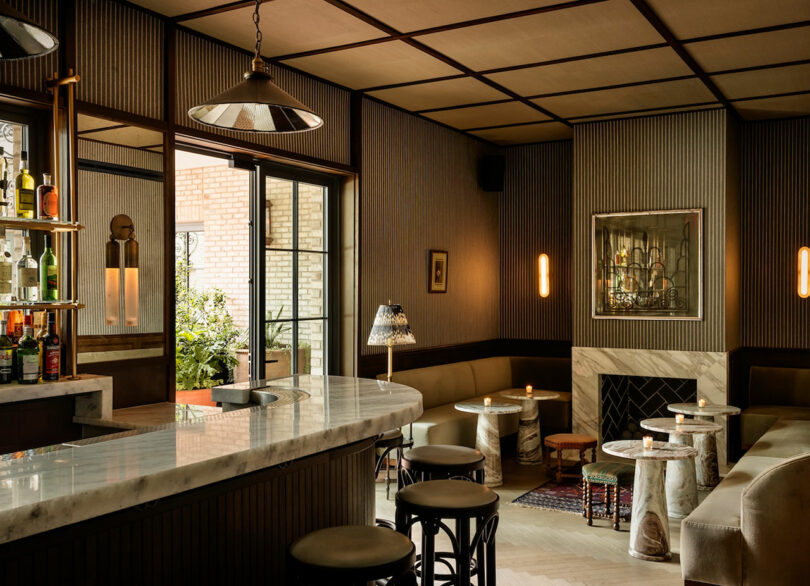 Elegant bar interior with a marble countertop, round tables, stools, plush seating, soft lighting, and a fireplace; large window shows greenery outside.