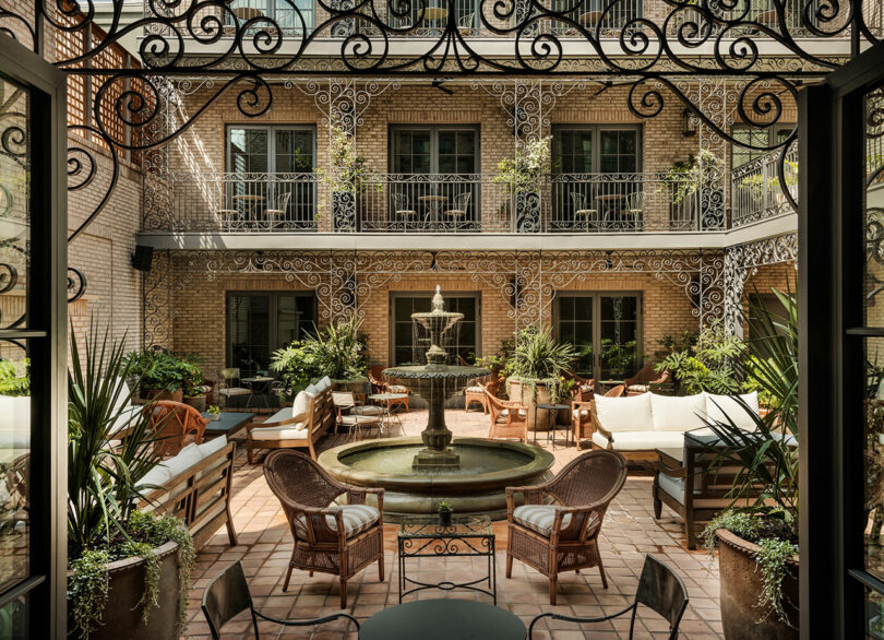 A courtyard with a central fountain, surrounded by wicker chairs, sofas, potted plants, and wrought iron balconies on a brick building.