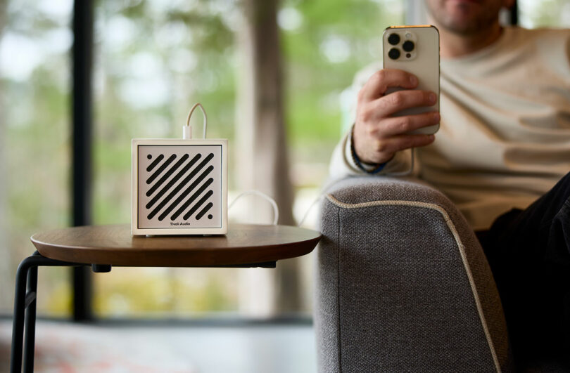 A person sitting on a sofa holds a smartphone, with a small white Tivoli speaker placed on a wooden side table in the foreground.