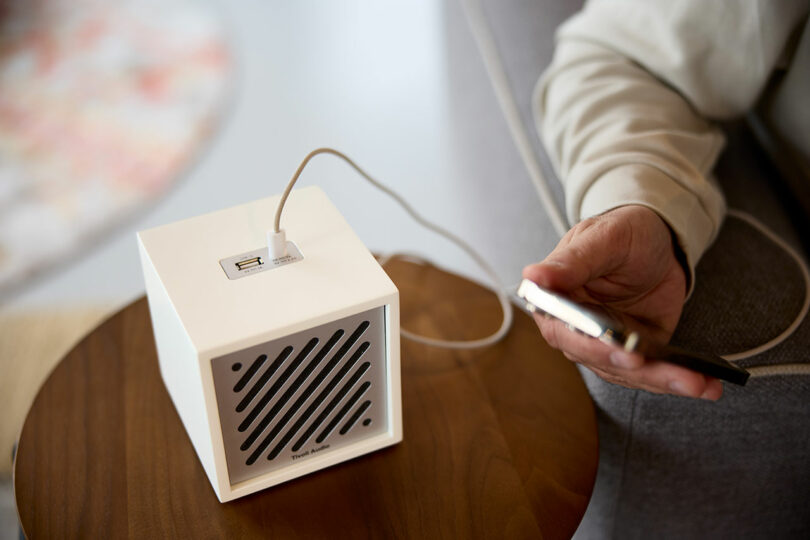 A person plugs a phone into a small, white, cube-shaped Tivoli device with a USB port, placed on a wooden table.