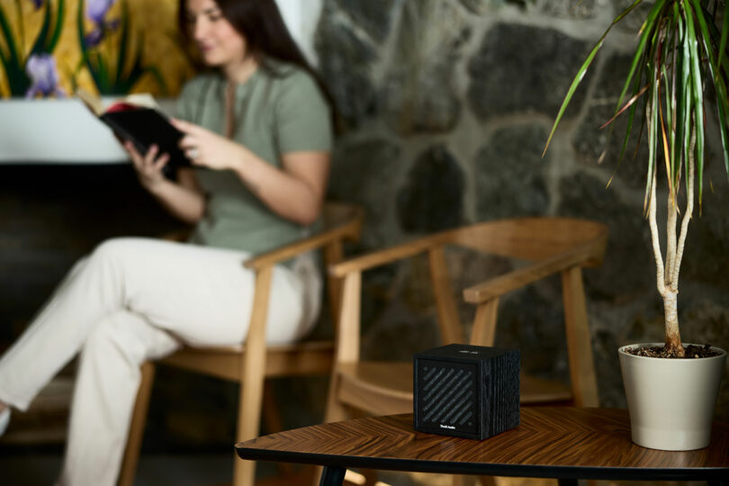 A small black Tivoli speaker sits on a wooden table next to a potted plant; in the background, a woman reads a book in a chair by a stone wall.