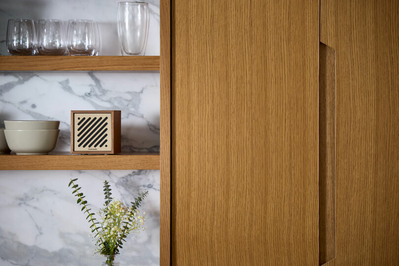 Wooden kitchen shelves with glasses, bowls, a Tivoli speaker, and a vase with flowers, set against a white marble backsplash next to a wooden cabinet.