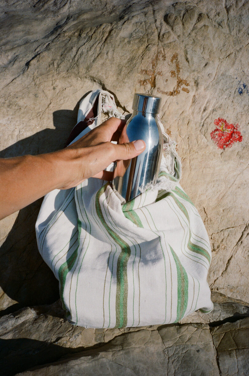 A hand holds an Aarke To-Go Bottle above a striped white and green cloth bag resting on a rocky surface.
