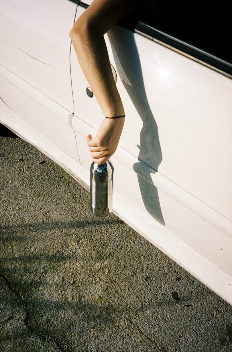 A person’s arm extends out of a car window, holding an Aarke To-Go Bottle above sunlit, cracked pavement.