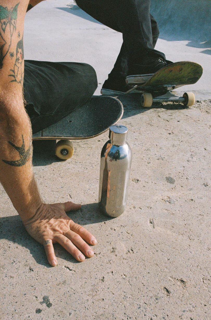 A person with tattoos kneels on pavement near a skateboard and an Aarke To-Go Bottle in a skatepark.