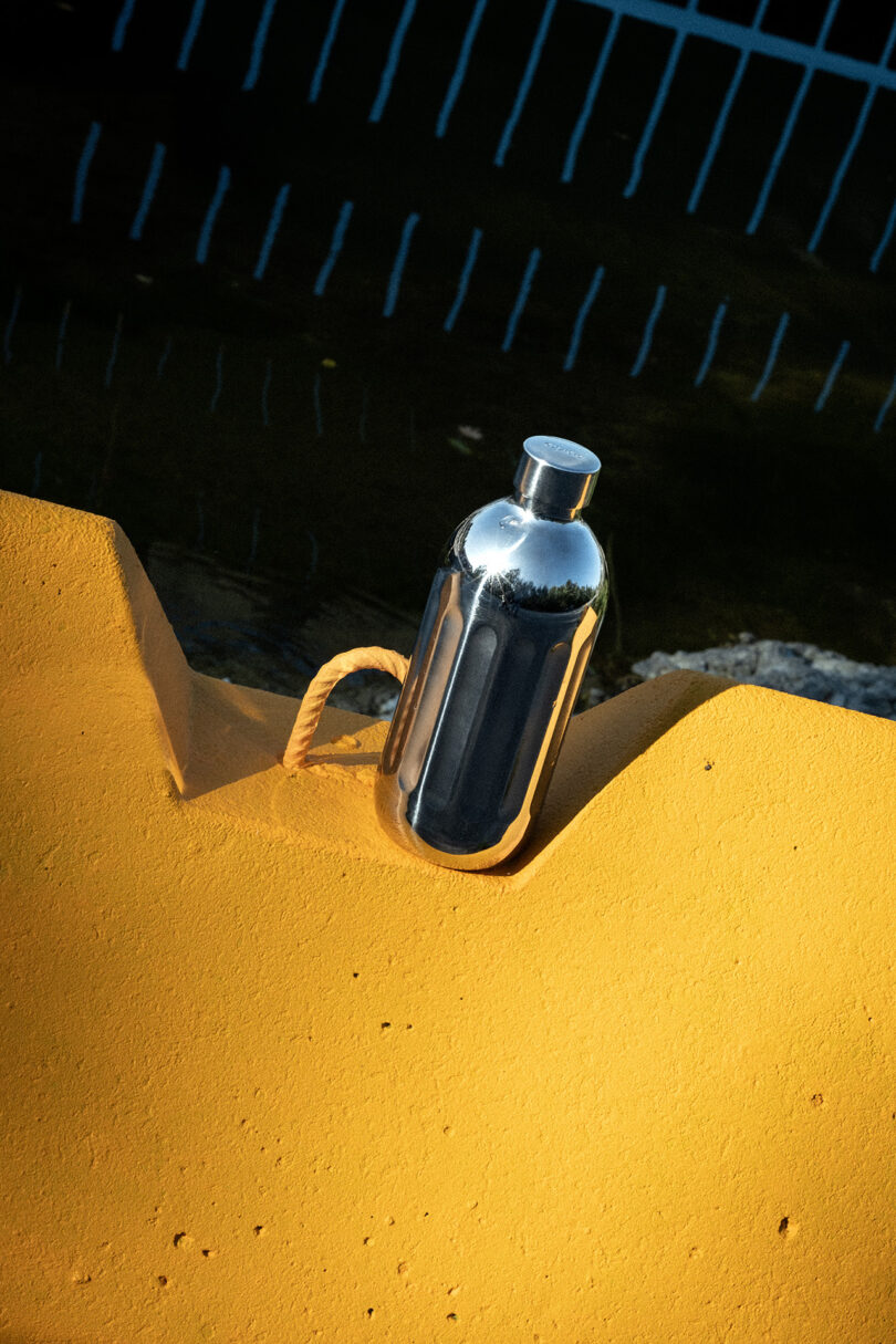A reflective To-Go Bottle rests on a yellow textured surface with a rope looped nearby; a body of water and blue fence are in the background.