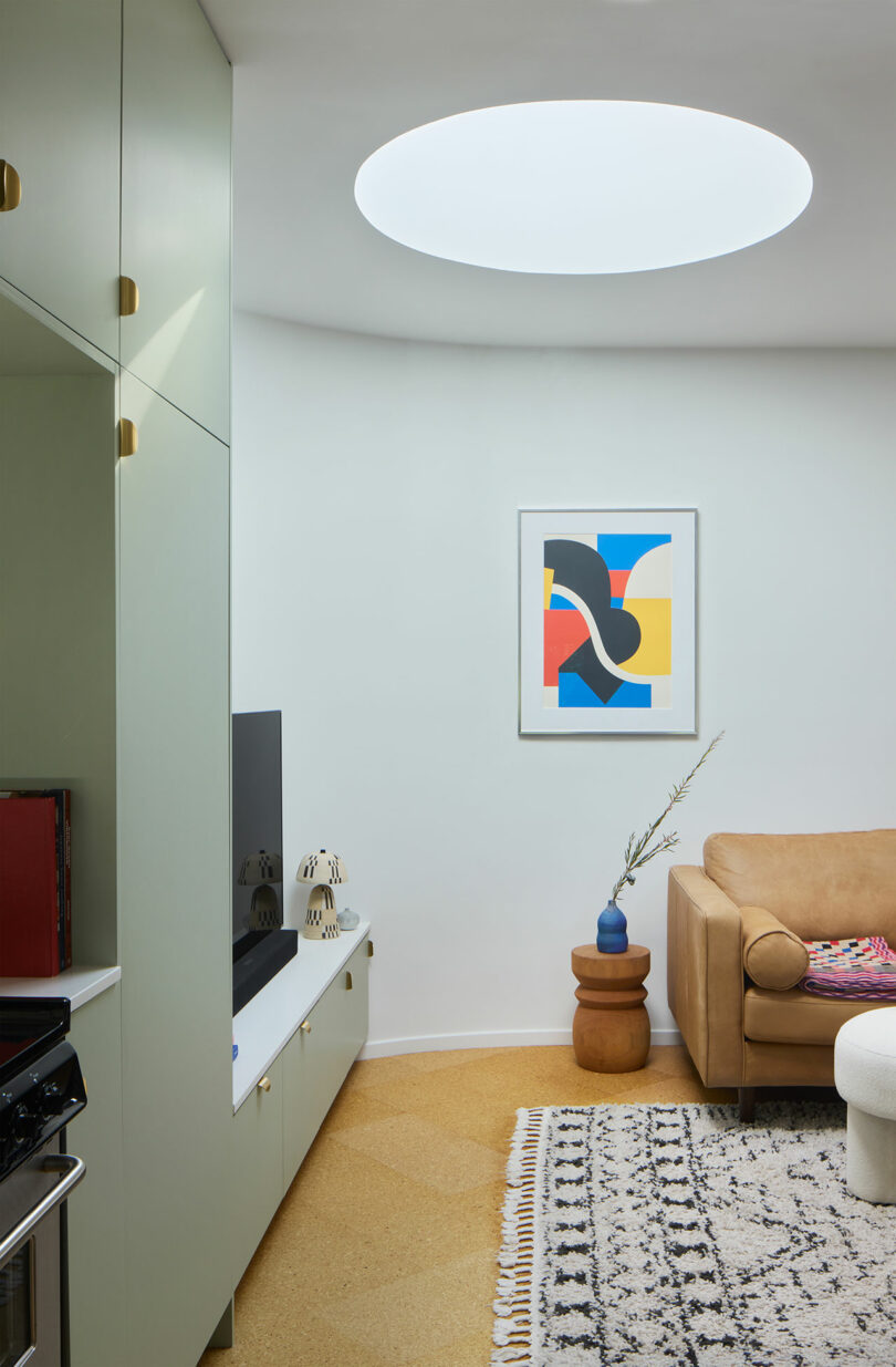A modern living room with a round skylight, abstract art on the wall, a beige sofa, a patterned rug, and a TV on a white cabinet.