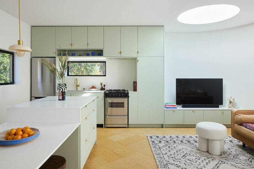 Modern kitchen and living area with pale green cabinets, stainless steel appliances, a wall-mounted TV, a round ottoman, and a bowl of oranges on a white island.