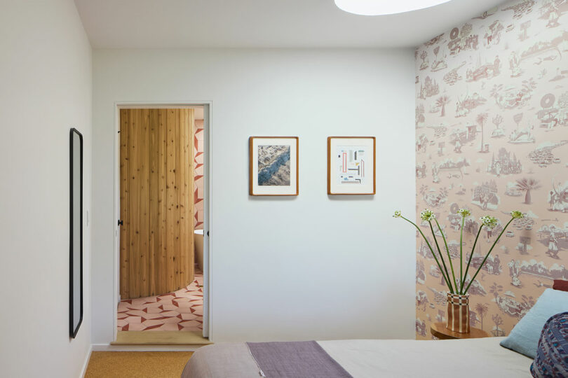 A modern bedroom with patterned wallpaper, a bed, a vase of flowers, two framed pictures on the wall, and an open doorway showing a wood-paneled room with geometric tile floor.