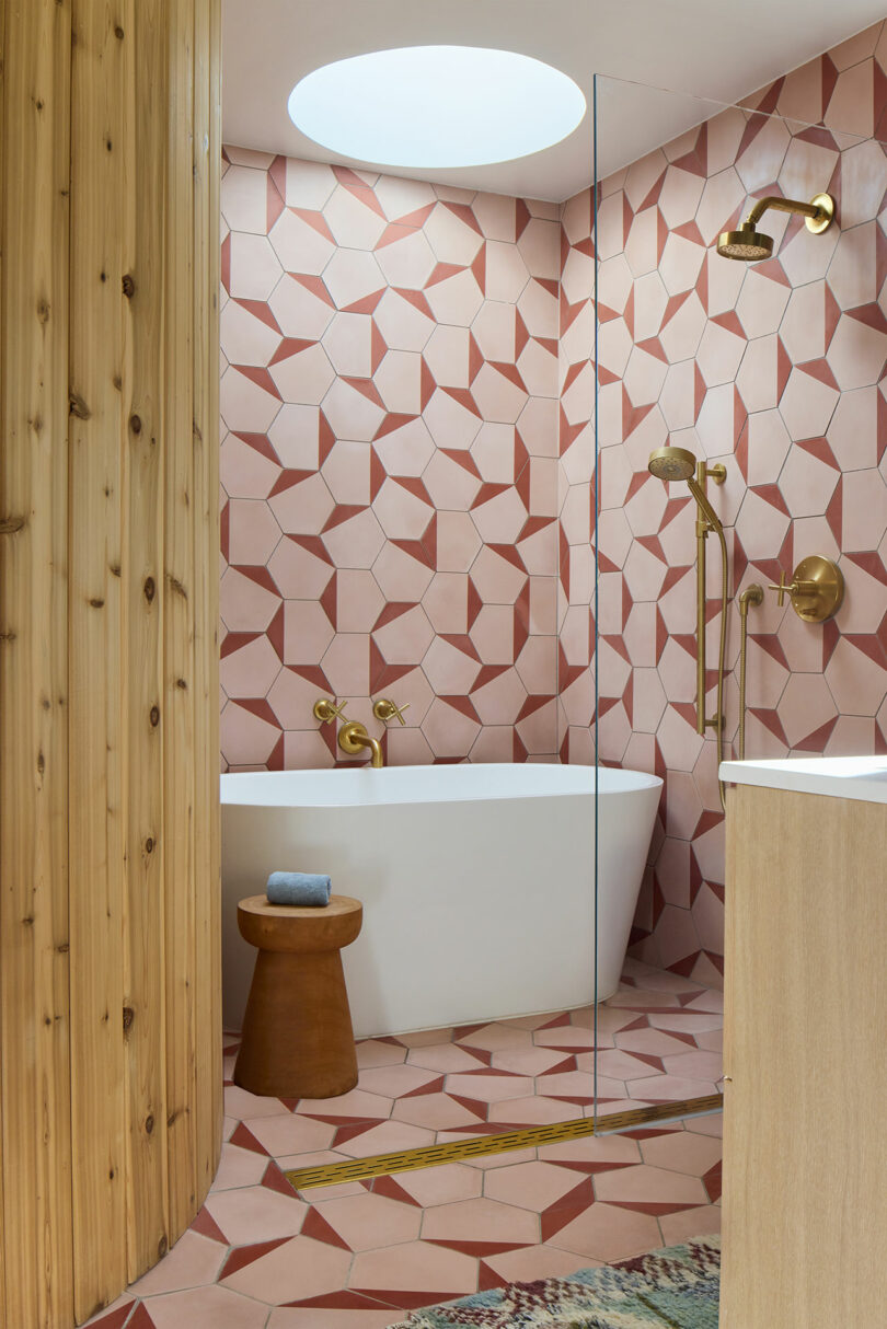 Modern bathroom with geometric pink tiles, a white freestanding tub, gold fixtures, a wooden stool, and natural wood accents under a round skylight.
