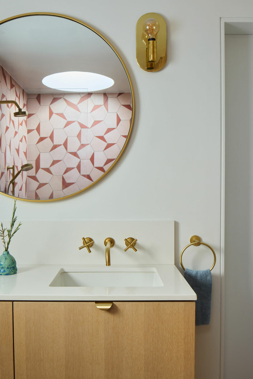 Modern bathroom with a round mirror, brass fixtures, light wood vanity, white countertop, and geometric pink tiles reflected from the shower.