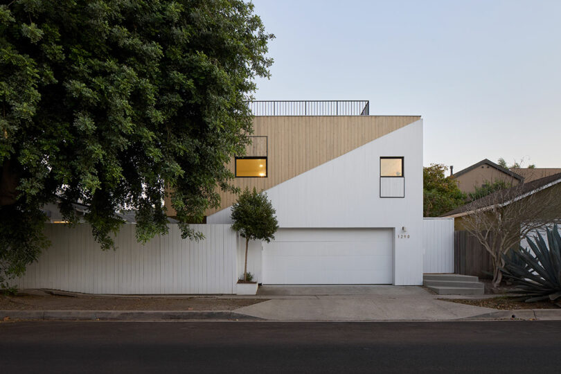 Modern two-story house with geometric facade, combining wood and white panels, a central garage, rectangular windows, and minimal landscaping.