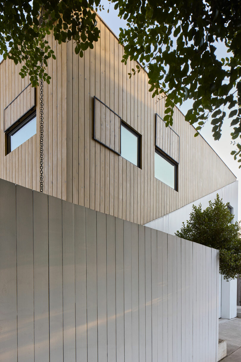 A modern building with light wooden siding and two rectangular windows, partially obscured by a tall white fence and tree branches at the top.