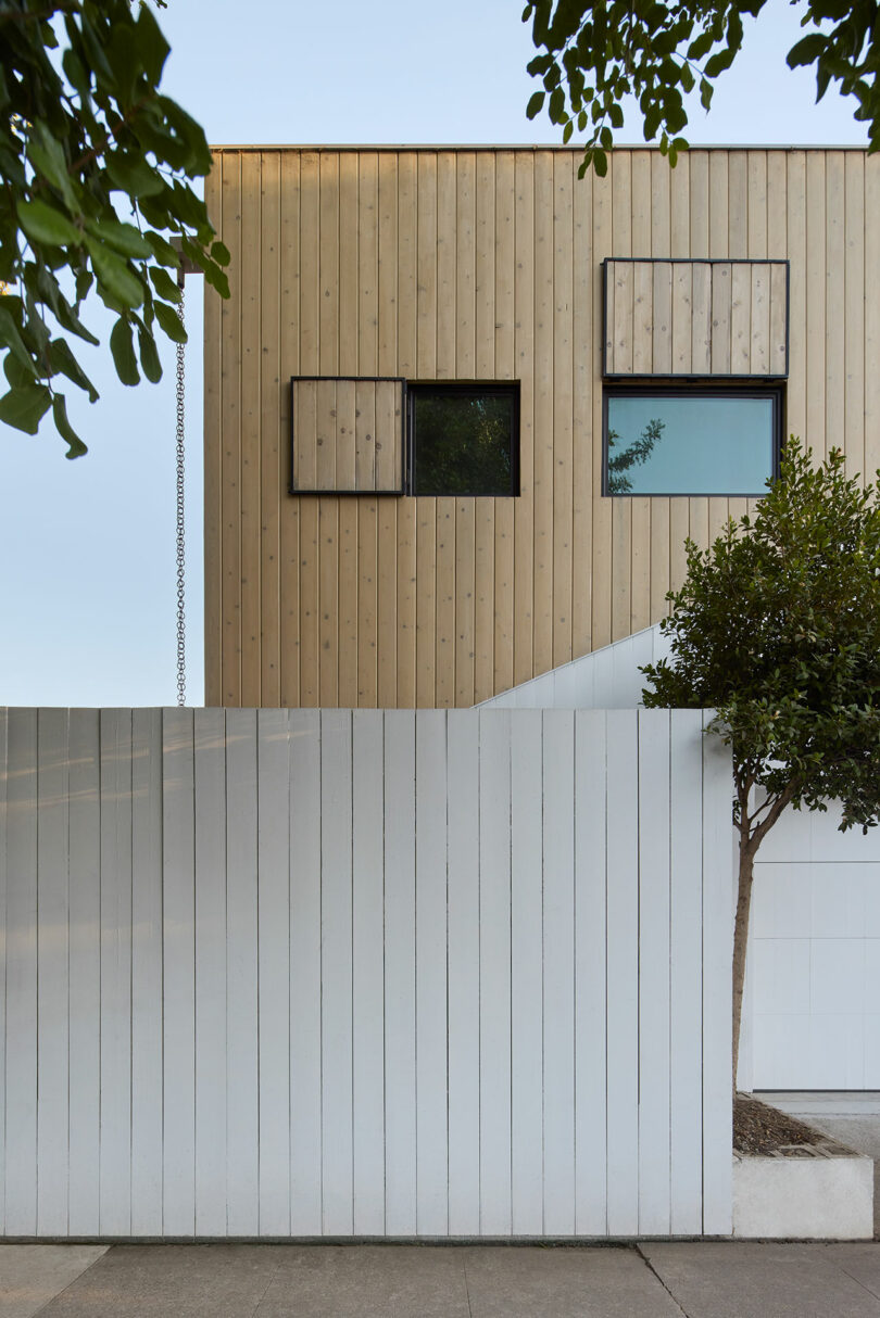 Modern building with light wood paneling features two rectangular windows, partially covered by square frames; white fence and tree in foreground.