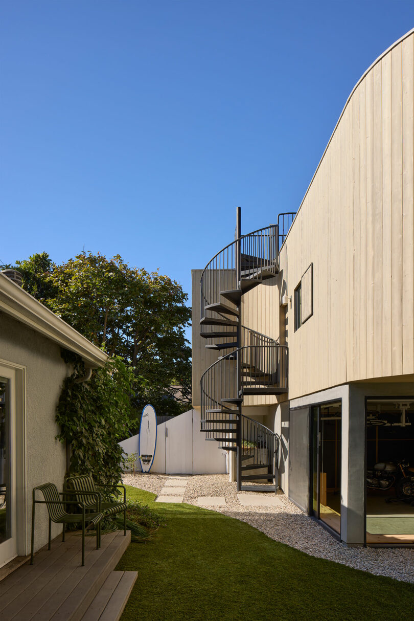 The modern building with a light wooden track contains an external spiral staircase, next to lawns and other structures, under a clean blue sky.