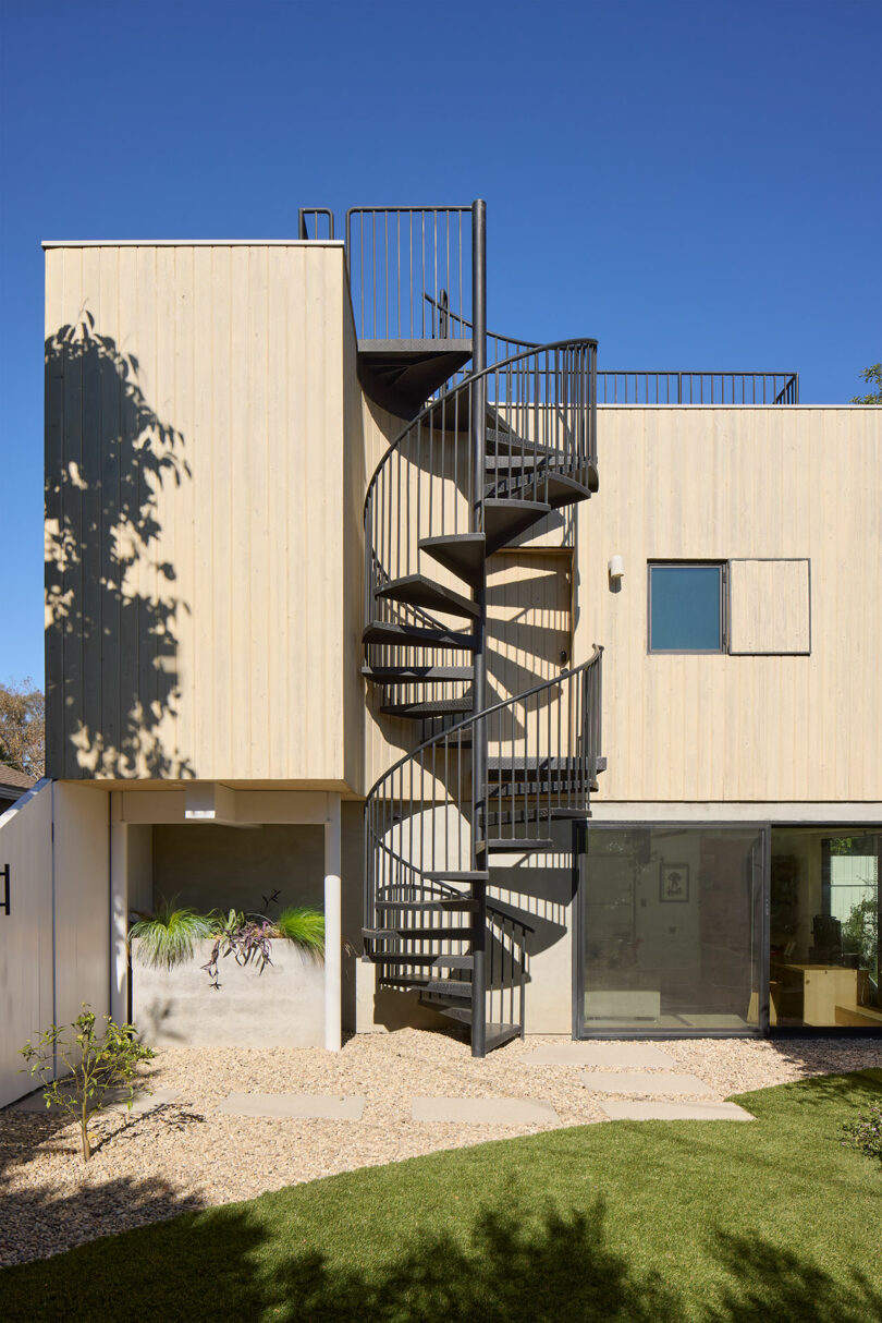 A modern two-story building with a light wood exterior features a black metal spiral staircase leading to a rooftop deck, set in a landscaped yard with grass and gravel.