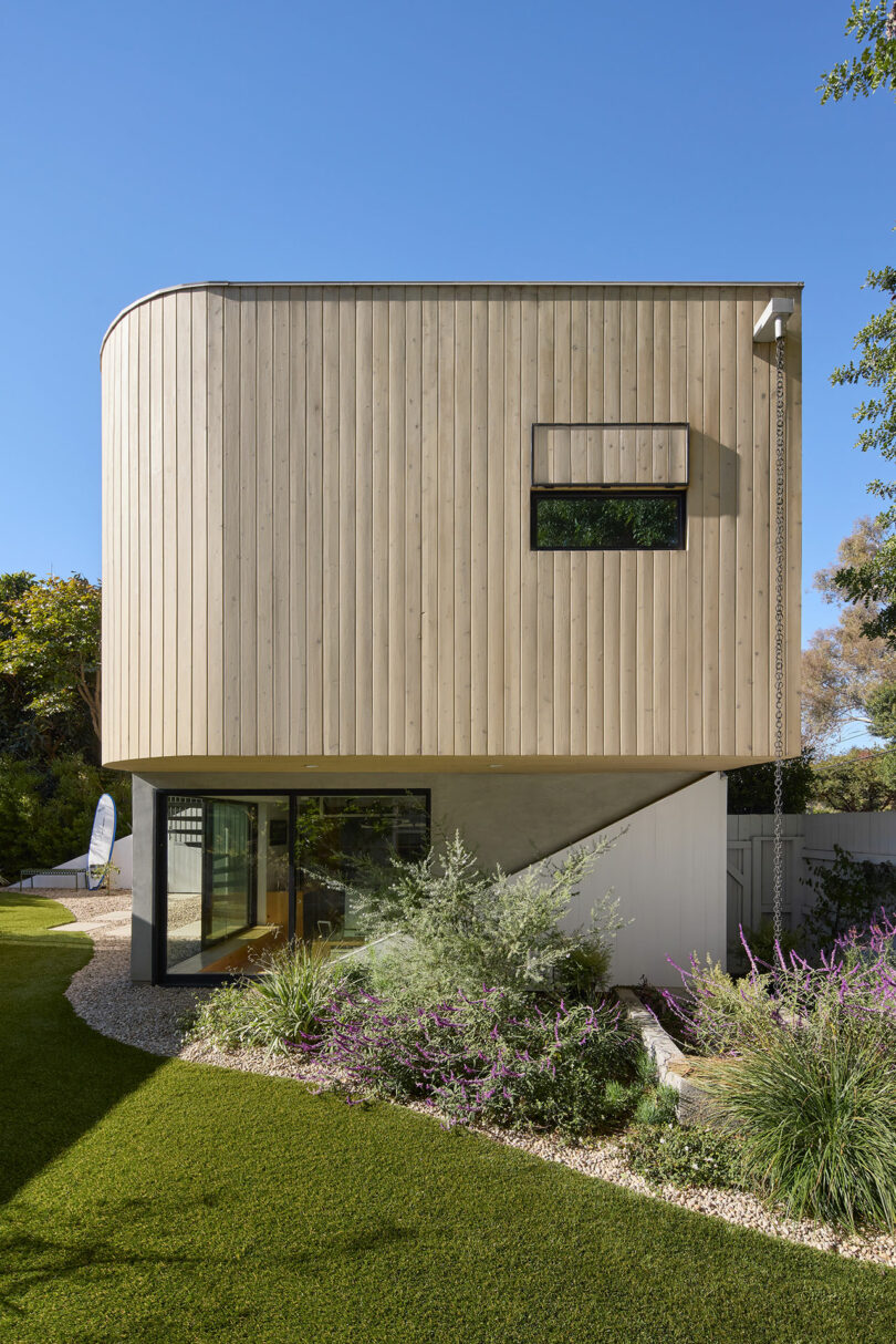 Modern two-story house with a curved wooden upper facade, large glass lower level, and landscaped garden with green grass and shrubs.