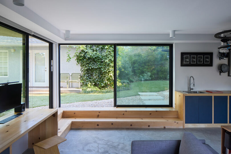 Modern living area with large sliding glass doors overlooking a green backyard, featuring wooden benches, a desk, a small kitchenette, and minimalist decor.