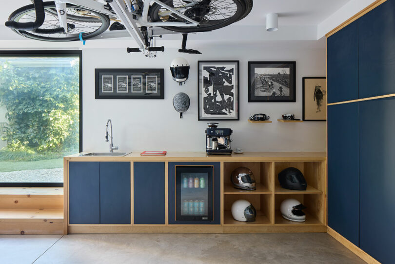 Modern room with blue and wood cabinets, a small sink, coffee machine, mini fridge, bike helmets, and bicycle mounted on ceiling; framed art hangs on the wall.