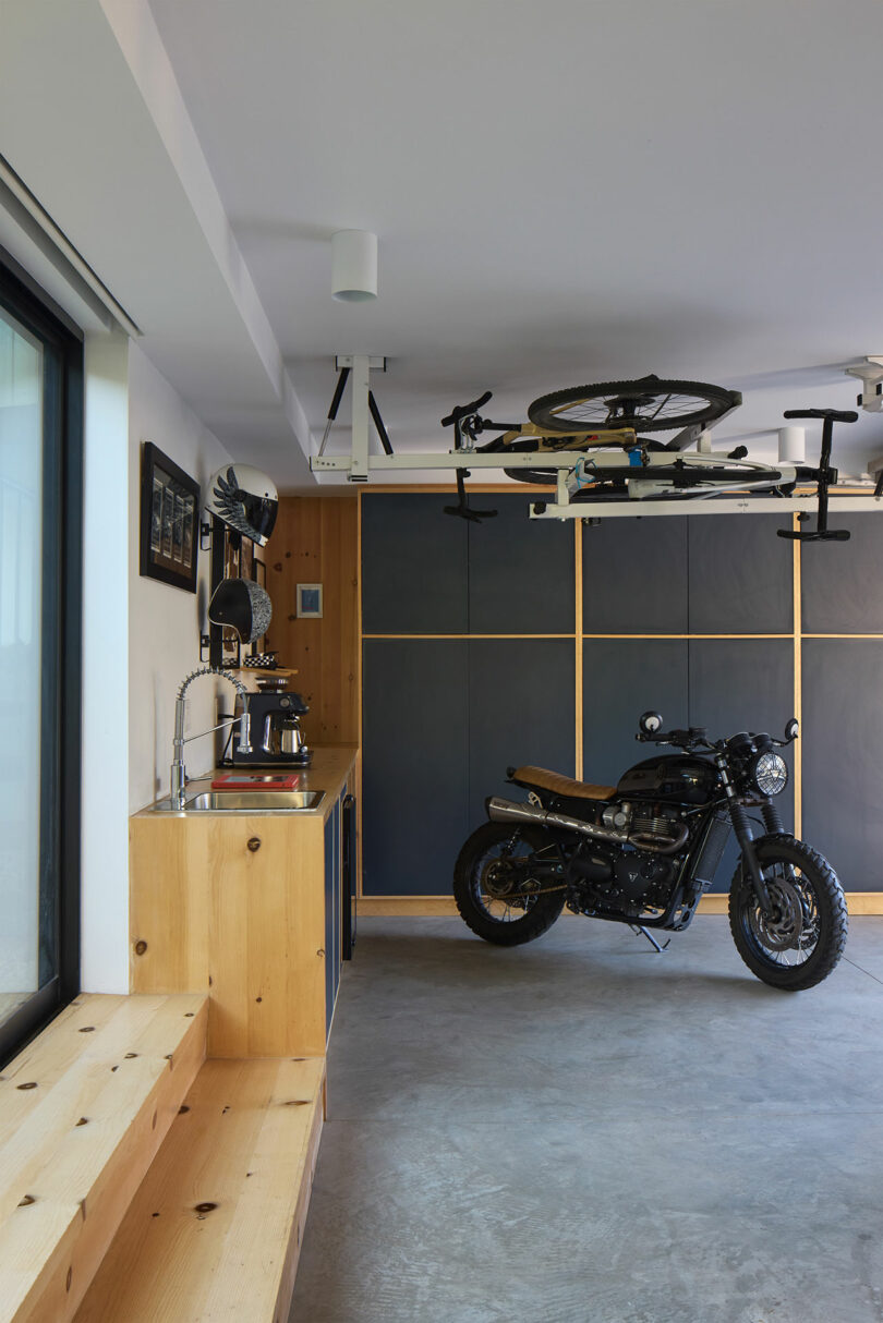 A modern garage interior with a motorcycle parked on a concrete floor, a bicycle mounted overhead, wooden cabinetry, and a countertop with a sink and coffee machine.