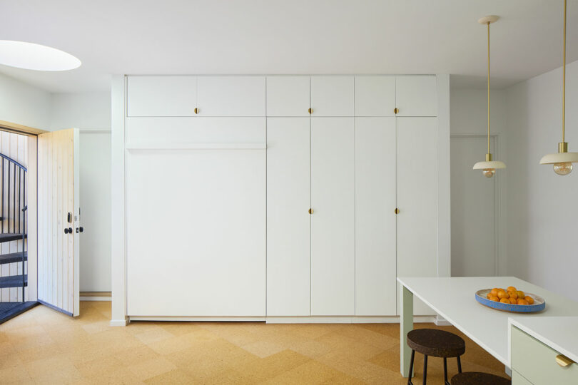 Minimalist kitchen with white built-in cabinets, a table with stools, pendant lights, and a bowl of oranges on the table. A door with a security gate is open on the left.