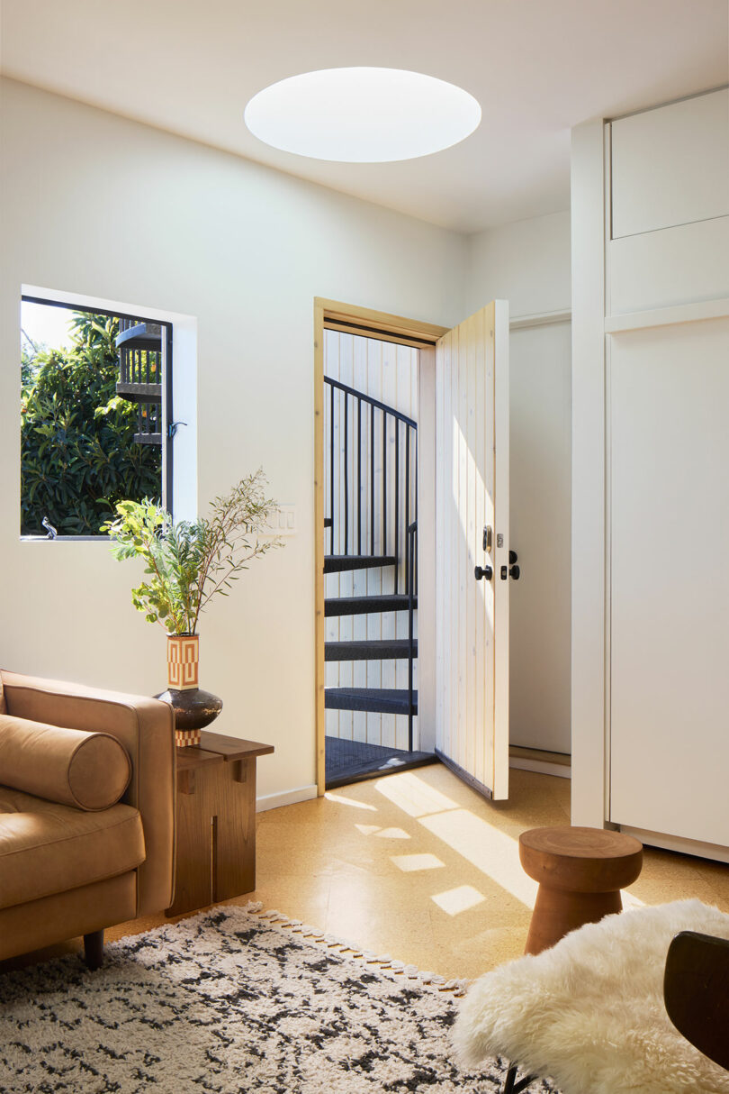 A modern living room with a tan sofa, patterned rug, and wood accents; the open door reveals an outdoor spiral staircase and greenery.